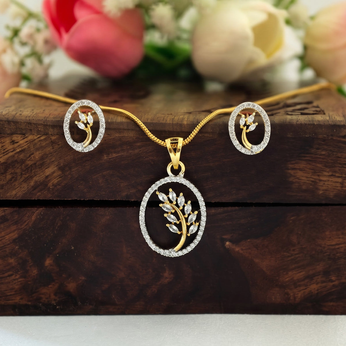 Gold leaf pendant necklace with matching stud earrings, displayed on a dark wooden box with blurred pink and white flowers in the background.