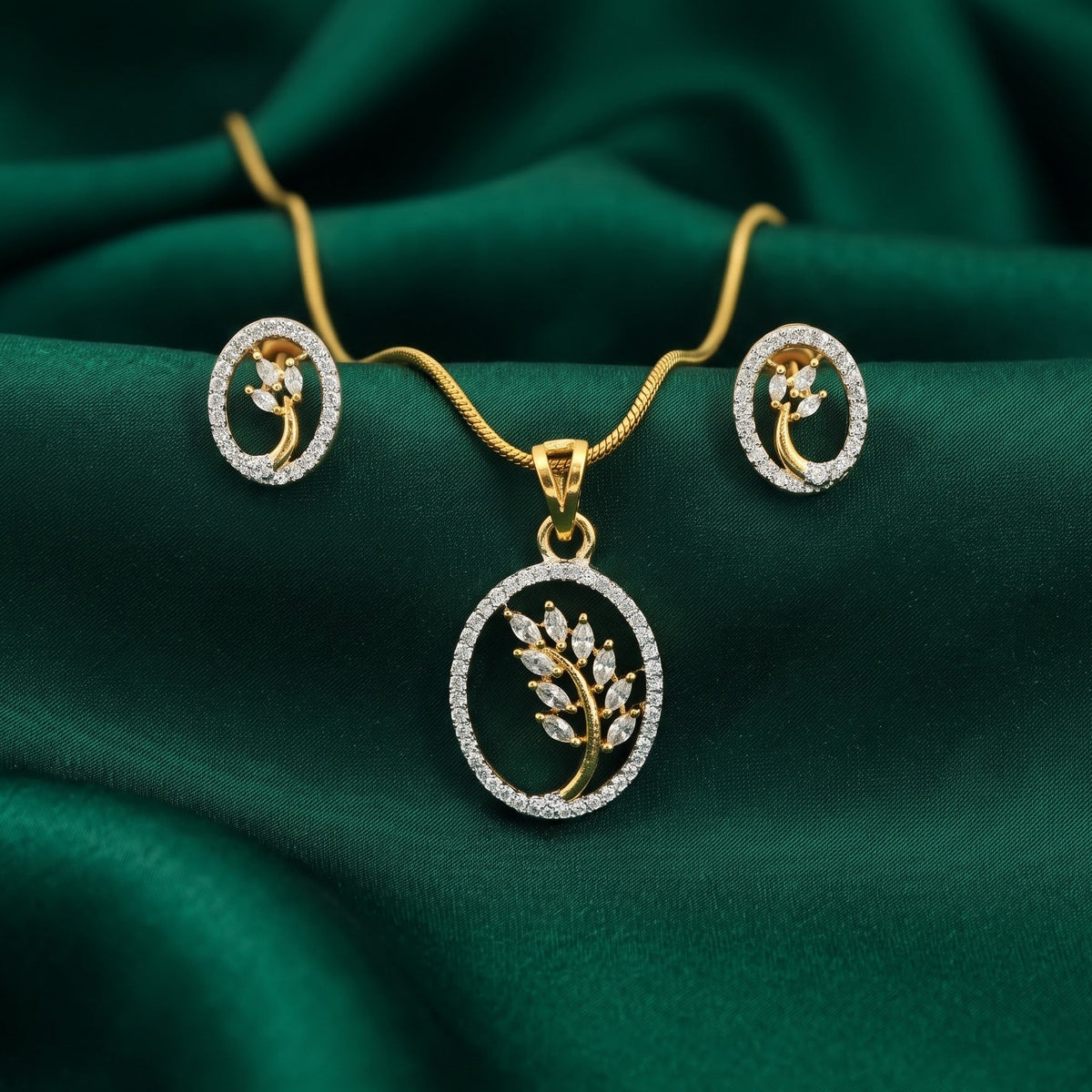 Gold jewelry set with a pendant necklace and matching earrings, featuring a leaf motif set in a circular frame with clear stones, displayed on a draped dark green satin fabric.