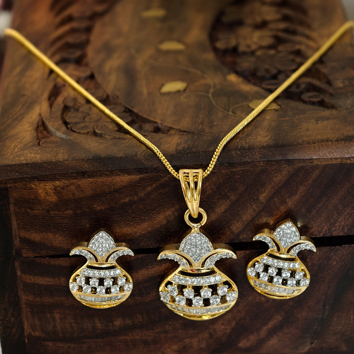 Gold-tone jewelry set with a pendant necklace and two matching earrings, each featuring a gold pouch-like charm set with clear stones, displayed on a carved wooden box with blurred pink and white flowers in the background.
