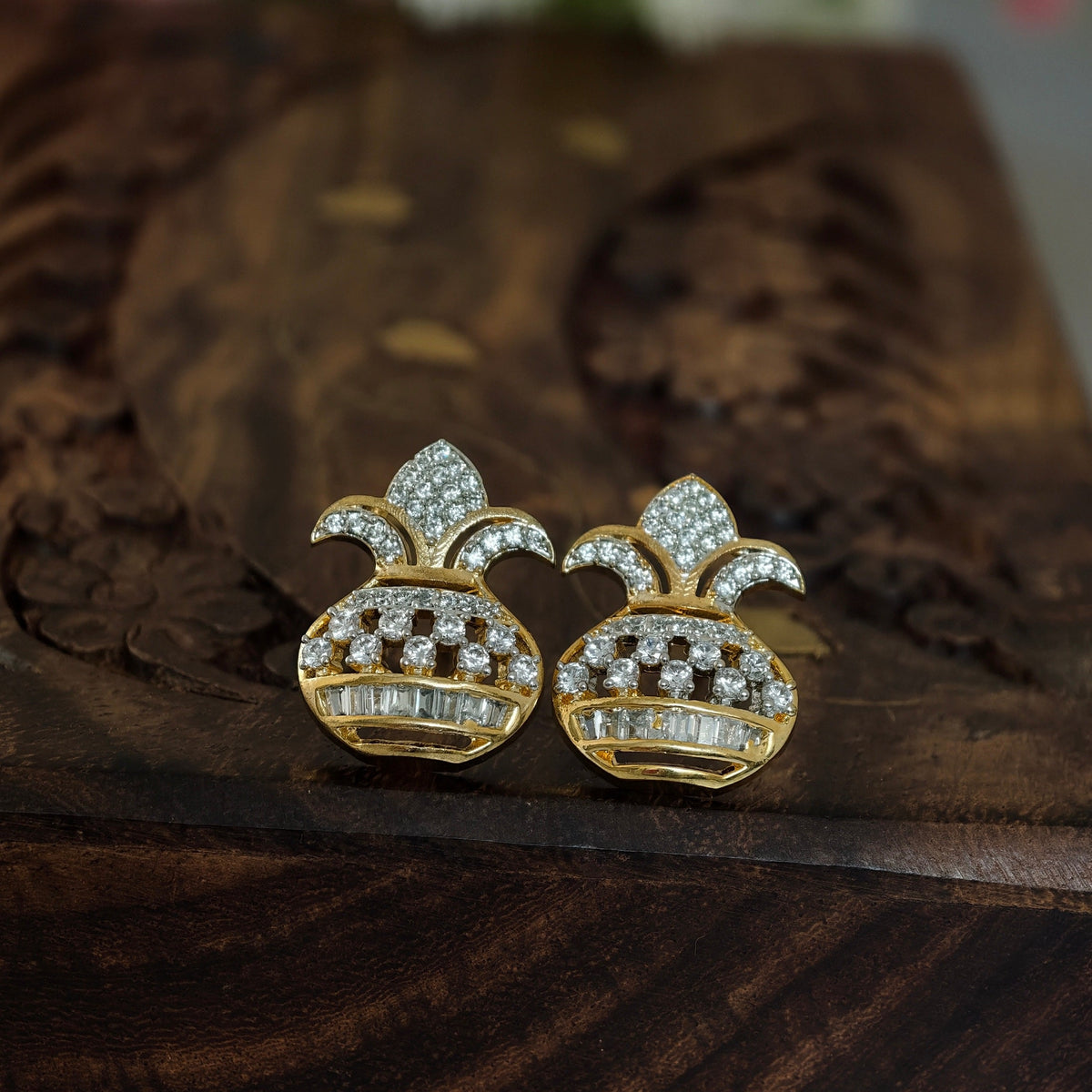 A pair of gold pineapple-shaped stud earrings paved with clear stones, resting on a dark wooden surface with a blurred floral background.