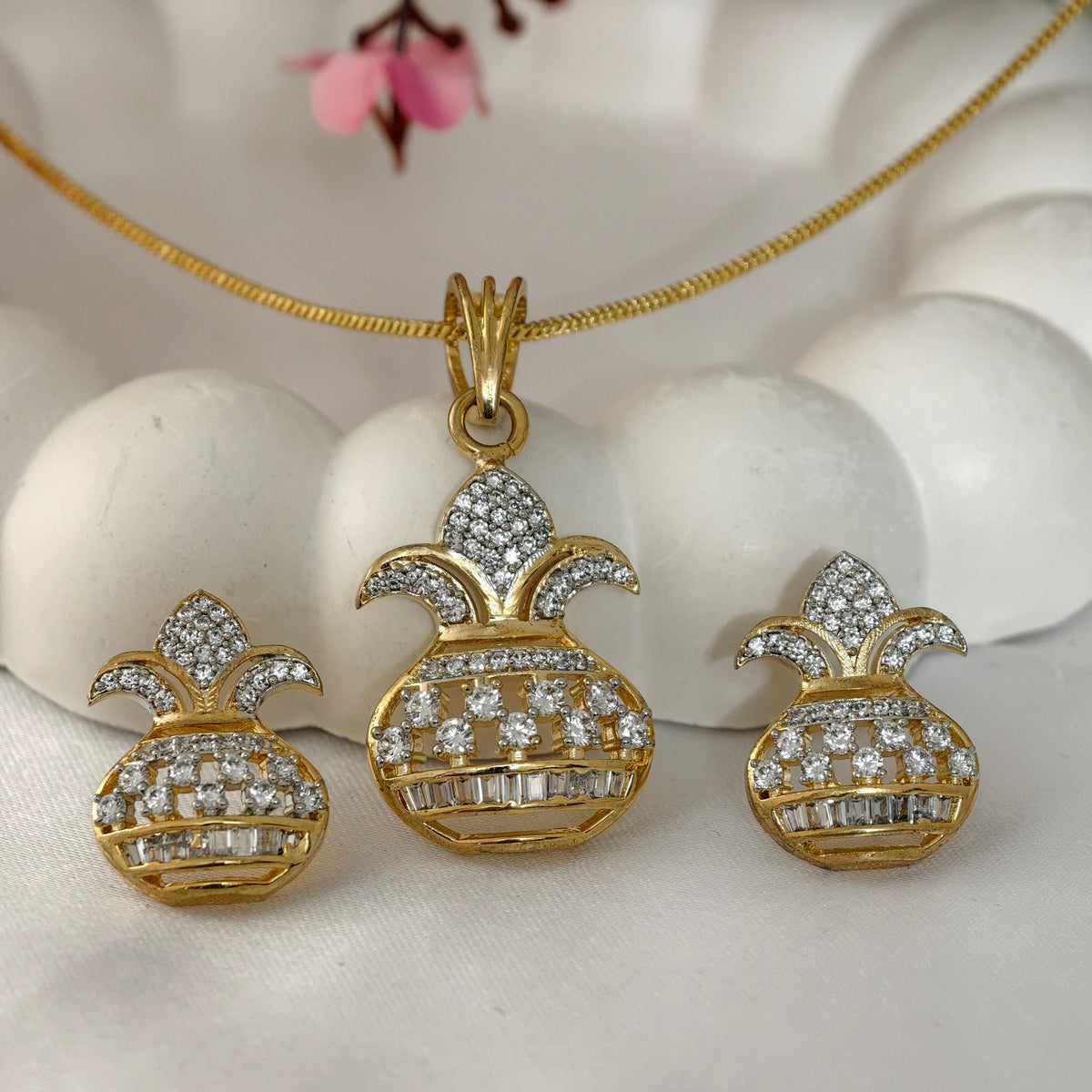 Gold pineapple pendant necklace with matching stud earrings, both adorned with clear pavé stones, displayed on a white textured surface with pink flowers in the background.