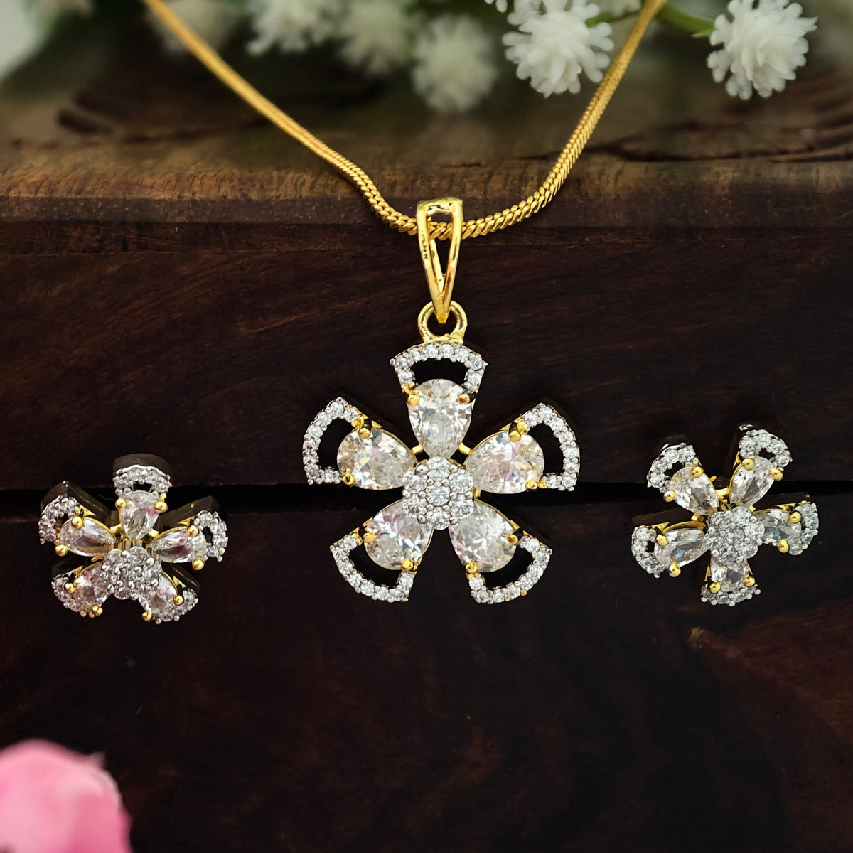 Gold-tone jewelry set featuring a cross-shaped pendant necklace, matching stud earrings, and a chain, decorated with clear rhinestones, displayed on a dark wooden surface with white and pink flowers.