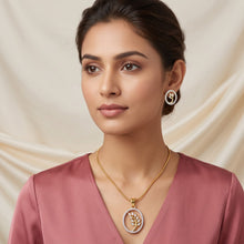 A woman wearing a gold circular pendant necklace with a white floral design and matching gold hoop earrings, paired with a dusty rose satin top, set against a draped cream fabric background.
