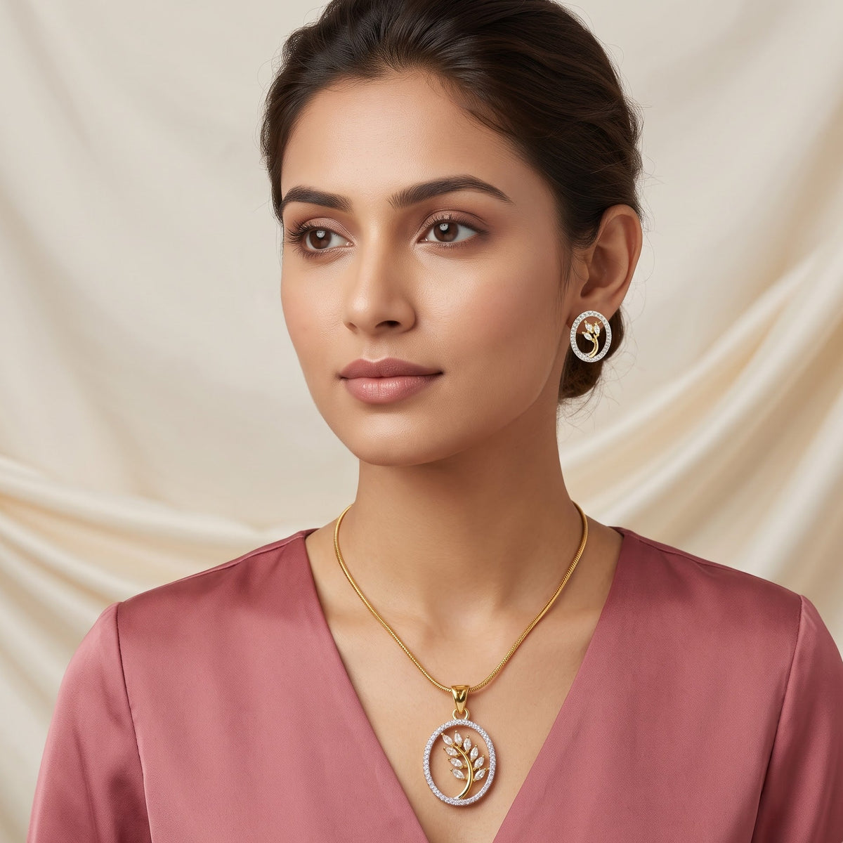 A woman wearing a gold circular pendant necklace with a white floral design and matching gold hoop earrings, paired with a dusty rose satin top, set against a draped cream fabric background.