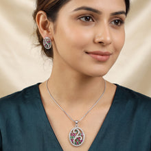 A woman wearing a silver-toned oval pendant necklace and matching stud earring. The jewelry features a central pink and green floral motif with a white circular background and a small red accent.